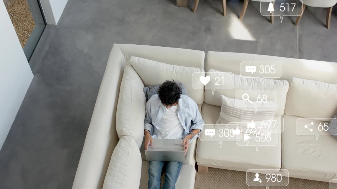 Man sitting on sofa using laptop, posting and receiving social media notifications flooding room
