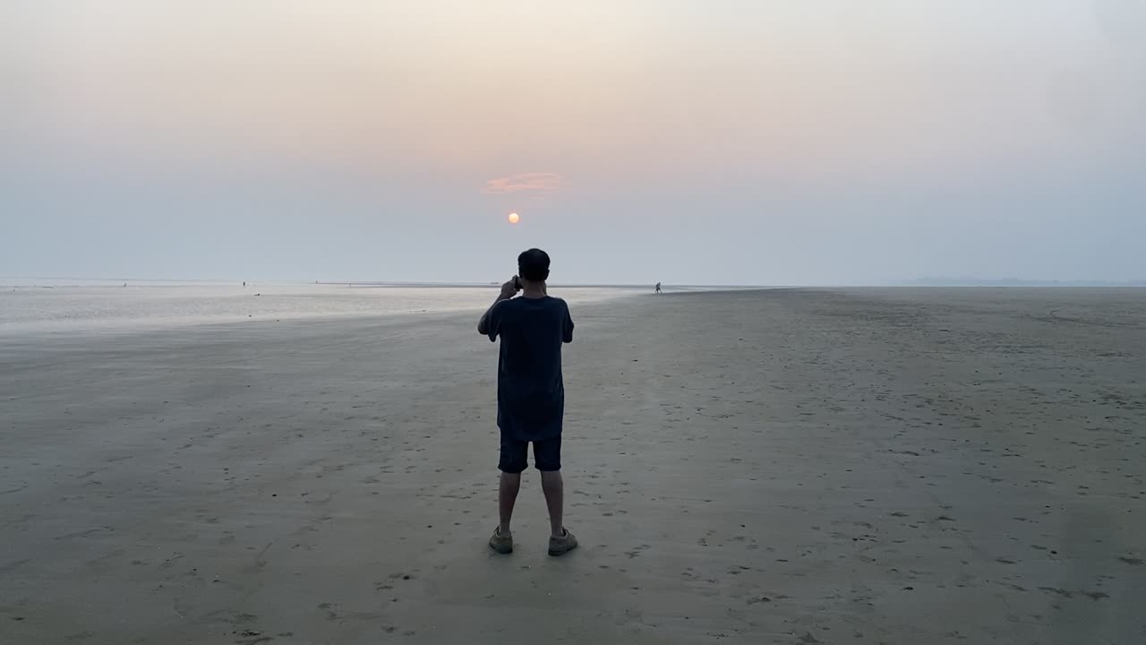 Rear cinematic shot of a man standing and clicking picture of the sunset at a lonely beach.