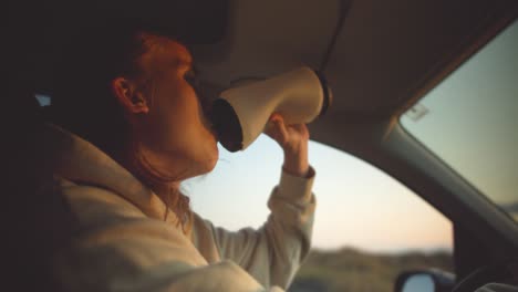 young blond man drinks from water bottle while driving in the light of the setting sun in the car