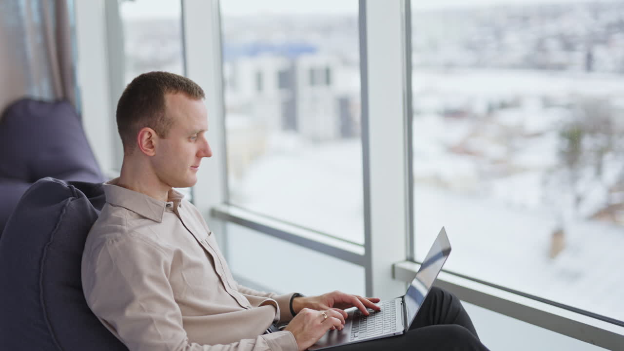 Calm man in bean bag chair works on laptop. Male sitting in front of panoramic window in spacious office. Blurred backdrop.