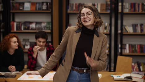 retrato de una mujer europea alegre y emocionada con chaqueta y vaqueros realizando una danza expresiva mientras escucha música en auriculares en la biblioteca académica contra estantes de libros de fondo y compañeros de clase alrededor