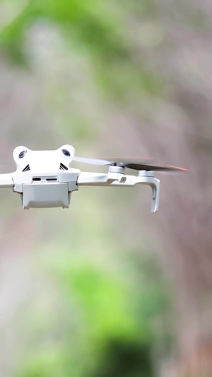 Drone quadricottero bianco si libra stabilmente all'aperto, circondato da fogliame verde, morbida luce naturale diurna, sfondo sfocato.