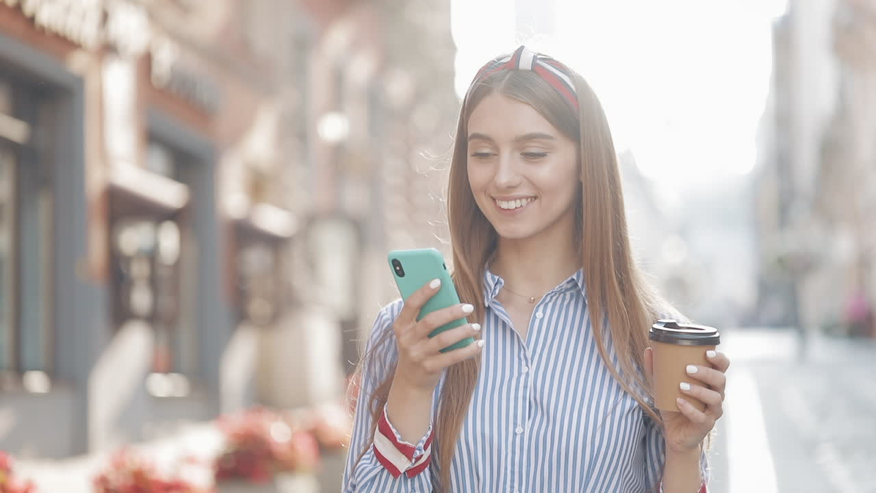Giovane donna beve caffè e sorride mentre usa lo smartphone per strada.