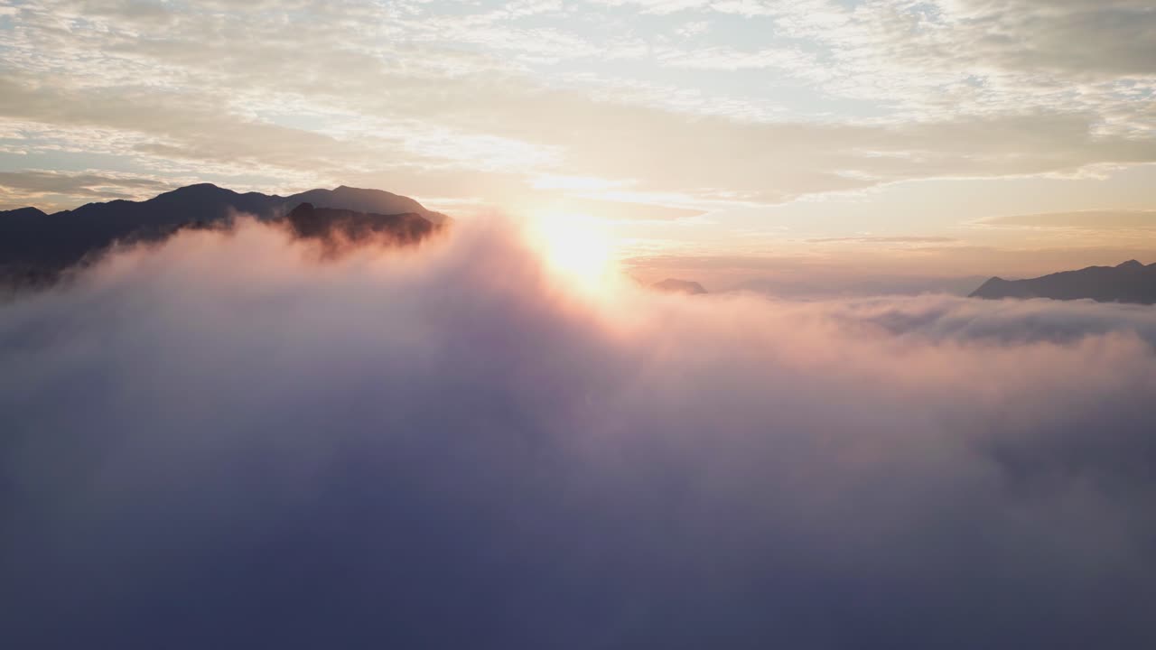 Veduta aerea con drone di aspre montagne che si ergono sopra una fitta coltre di nuvole durante un'alba colorata. Nuvole infinite riempiono valli profonde creando un effetto di mare di nuvole ad alta quota