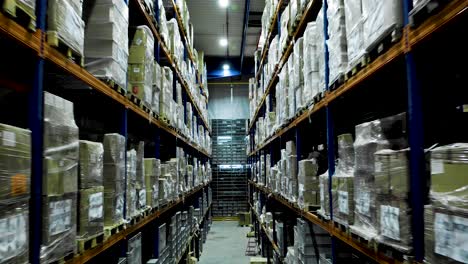 Drone-flies-between-rows-of-stacked-shelves-in-industrial-warehouse,-dolly-out