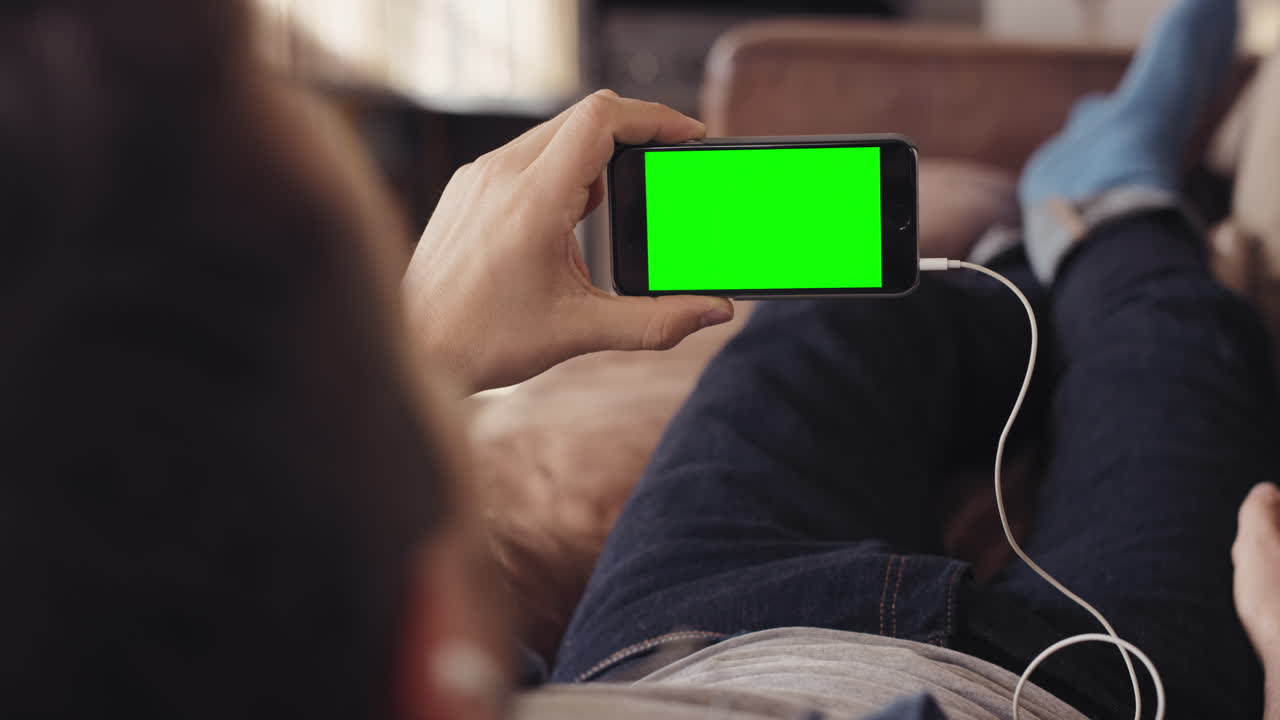 멋진 남자가 스마트 폰을 사용하여 집에서 소파에서 녹색 화면 비디오를보고 있습니다.