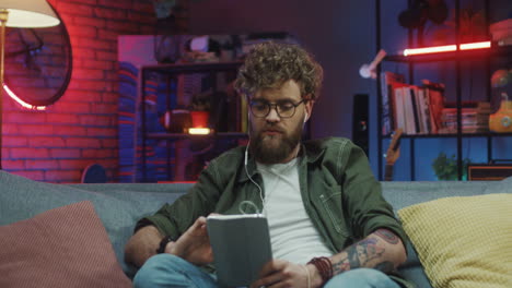 joven con gafas y auriculares sentado en el sofá con una tableta viendo algo o escuchando música por la noche