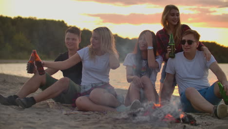 Die-Jungen-Leute-Sitzen-Um-Ein-Lagerfeuer-Am-Flussufer.-Sie-Lachen-Und-Singen-Lieder-Bei-Bier-Bei-Sonnenuntergang-An-Einem-Sommerabend.