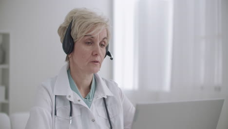 aged female family doctor is consulting online talking with patient by video chat at laptop working remotely