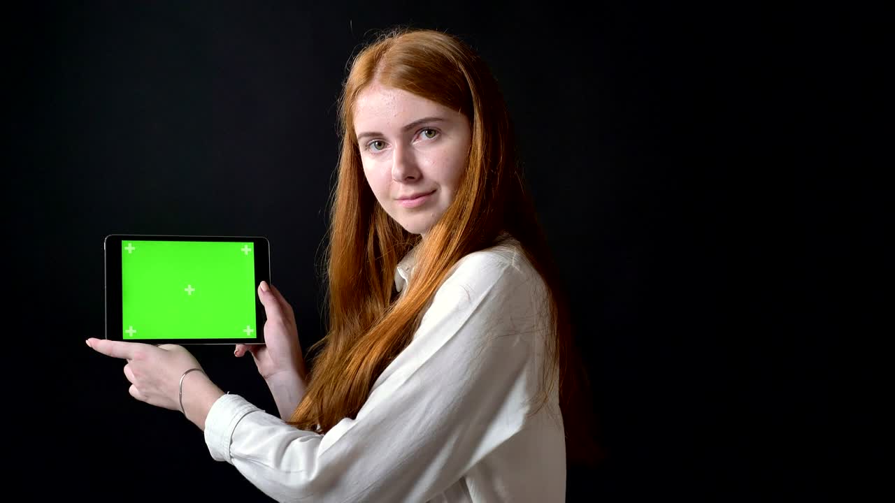 Ginger young woman showing thumbs up to tablet with chromakey and nodding, smiling, isolated on black background