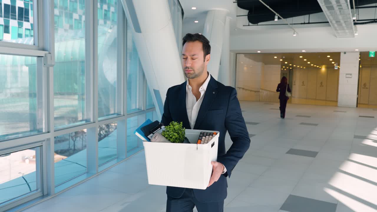 Empresário triste caminha pelo saguão moderno do escritório, segurando uma caixa branca com arquivos e uma planta, após demitir a empresária rival, que joga a planta de volta na caixa e se afasta celebrando.