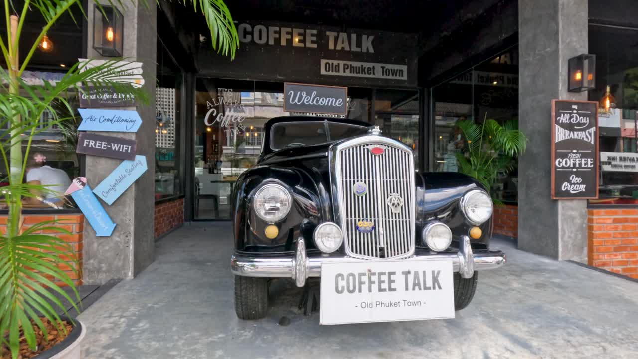 Camera slowly approaches vintage car at retro coffee shop entrance, daylight, urban Phuket, Thailand