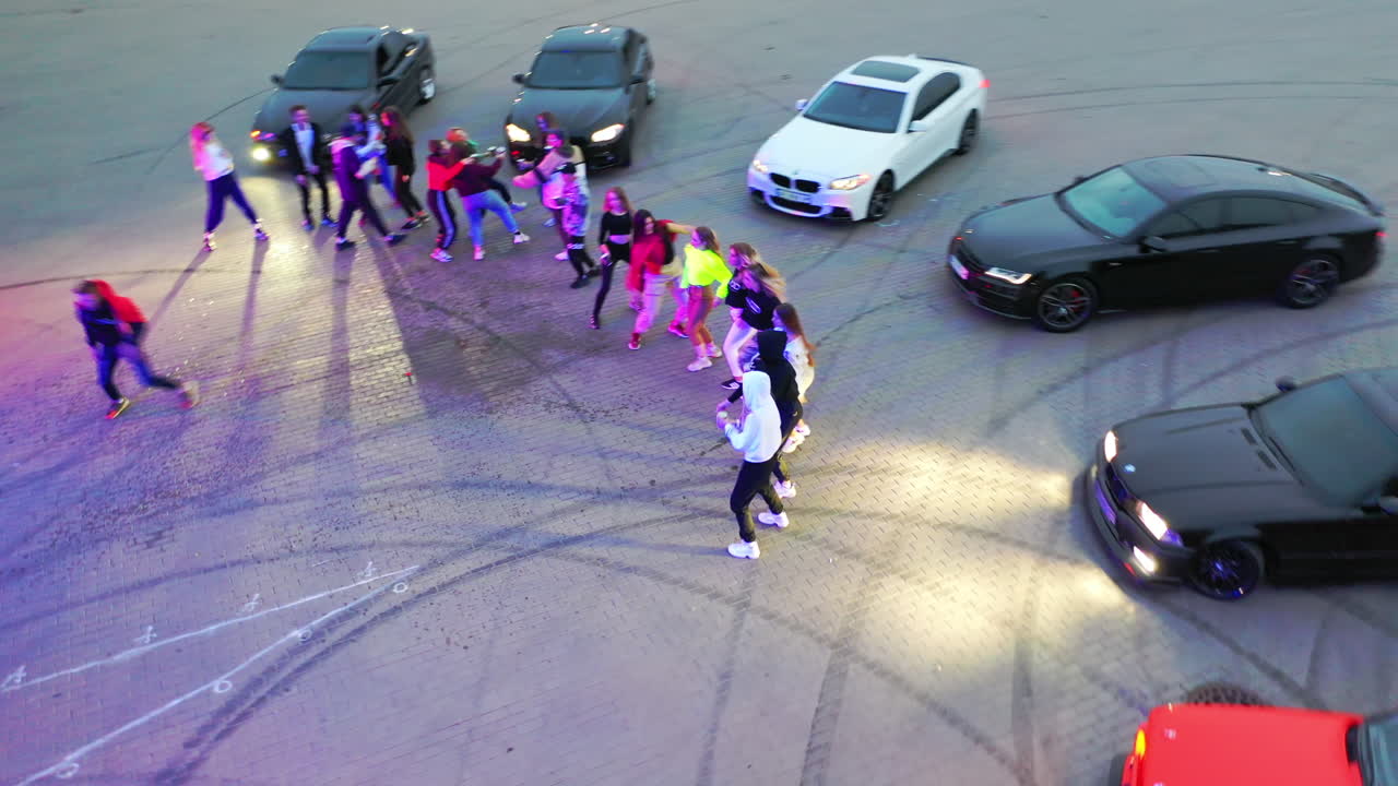 Party at the drifting field. Young happy people dancing near the race cars outdoors at dusk. Top view.