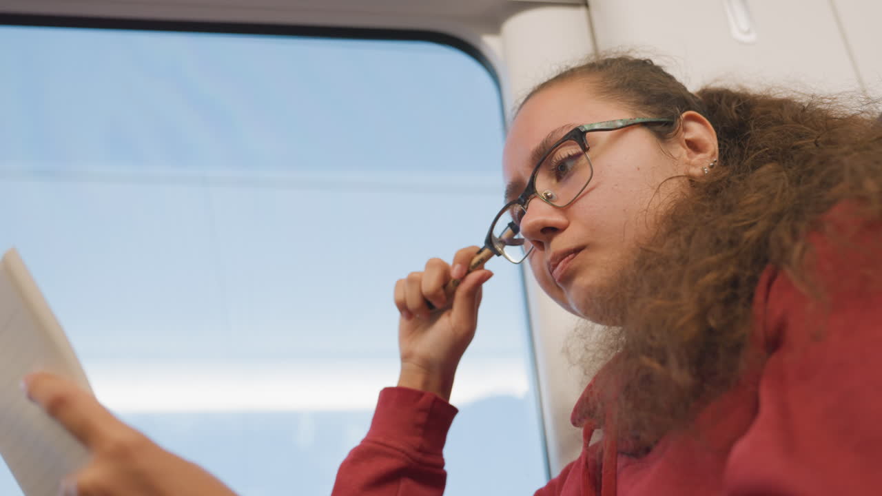 Young woman reading notes by train window, glasses lifted, pen in hand, pages turned, soft daylight, suburban scenery passing, itinerary planning, study pause, reflective commuter mood
