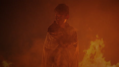 a young person wrapped in a cloak against a fiery, smoky background