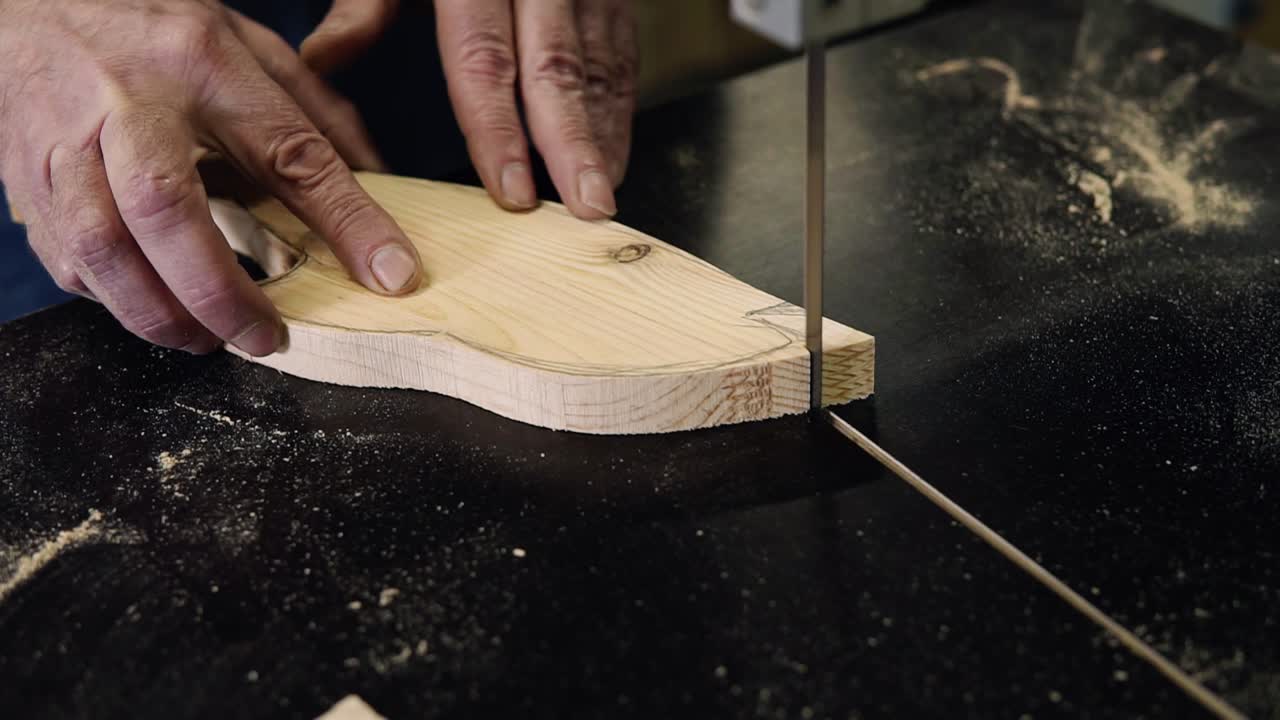 Close up footage of male's hands working with an electric cutting machine. High angle footage of a man cutting a fish shape wooden pattern on a table. Slow motion