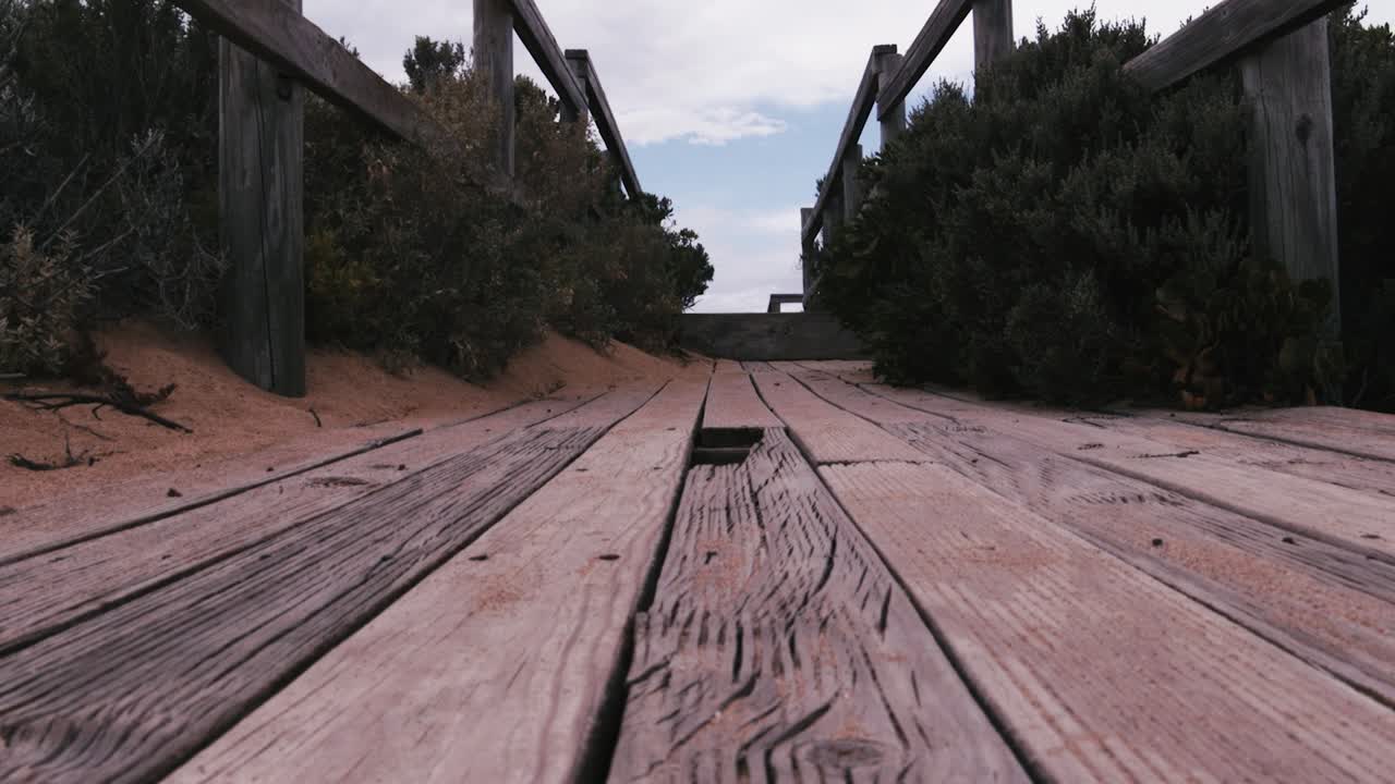 A weathered boardwalk stretches through lush coastal foliage under a cloudy sky