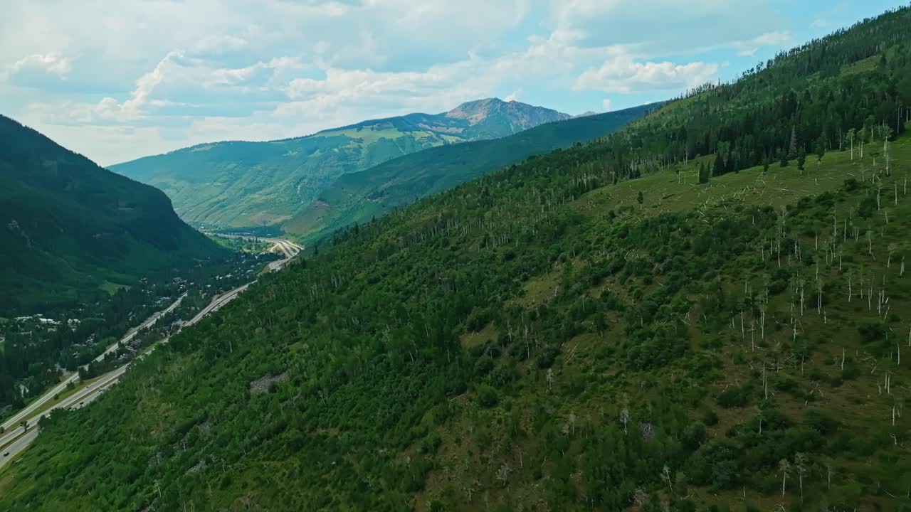 Drone tracking along Vail mountain ridge with deep green forest and blue summer sky above as cars drive on highway below