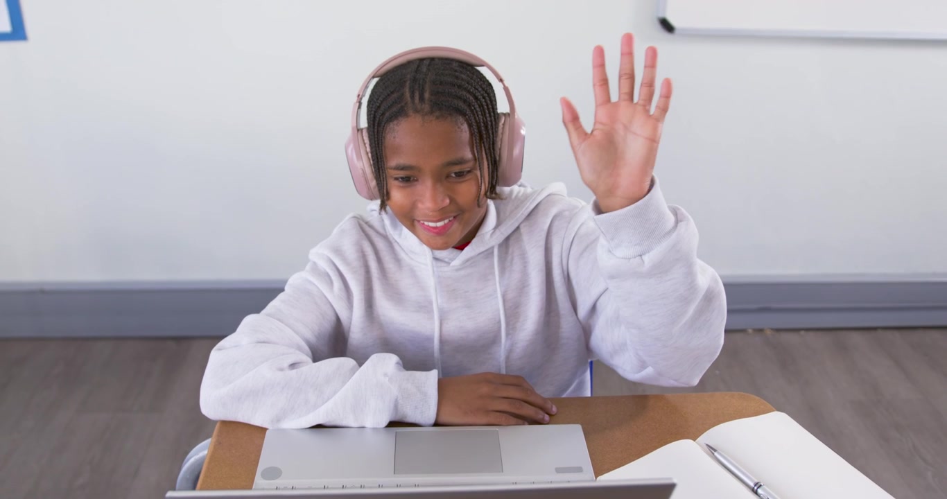 Adolescente afro-americana participando de uma chamada online para a aula, sorrindo, acenando, ajustando o laptop e o fone de ouvido.