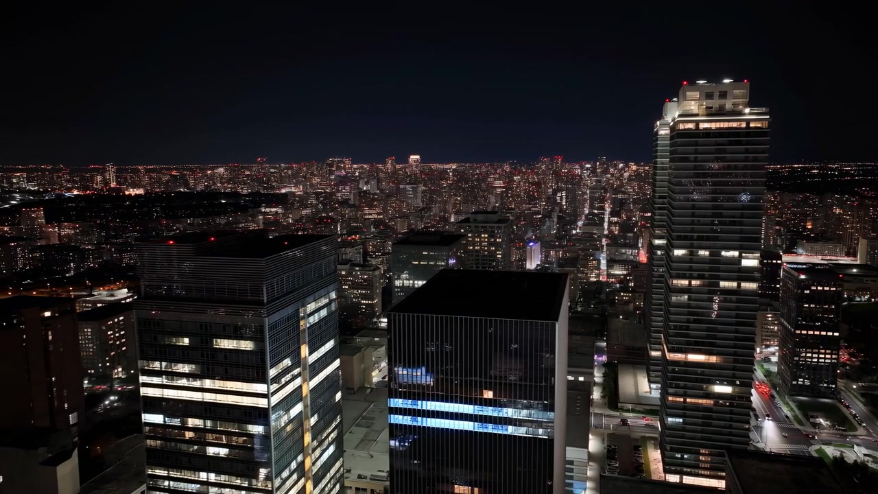 Vista noturna do denso horizonte do centro da cidade com janelas de escritórios brilhantes, torres de vidro reflexivas, blocos residenciais mistos e sutis rastros de luz em movimento serpenteando pelas ruas abaixo.