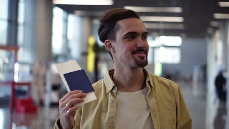 happy, handsome bearded american man showing passport with flight tickets and waving it. looking and smiling to the camera