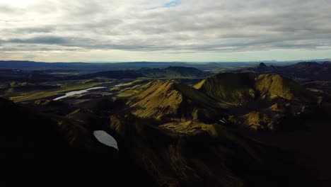 Aerial-landscape-view-over-Icelandic-highlands,-with-dark-hills-and-mountains,-covered-in-bright-green-grass,-on-a-cloudy-day