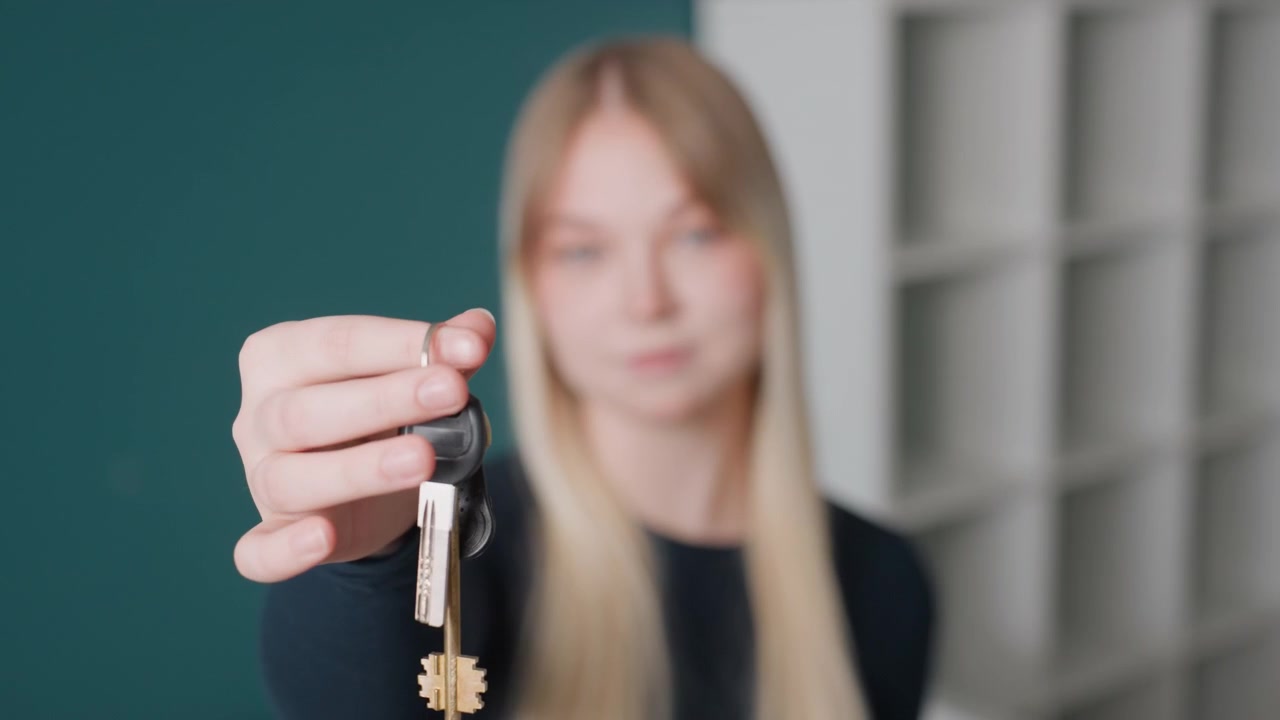 Donna caucasica presenta le chiavi in primo piano in un appartamento non arredato con sfondo verde acqua e scaffalature, donna bionda con espressione calma, mano ferma che estende il portachiavi verso la fotocamera, consegna professionale.