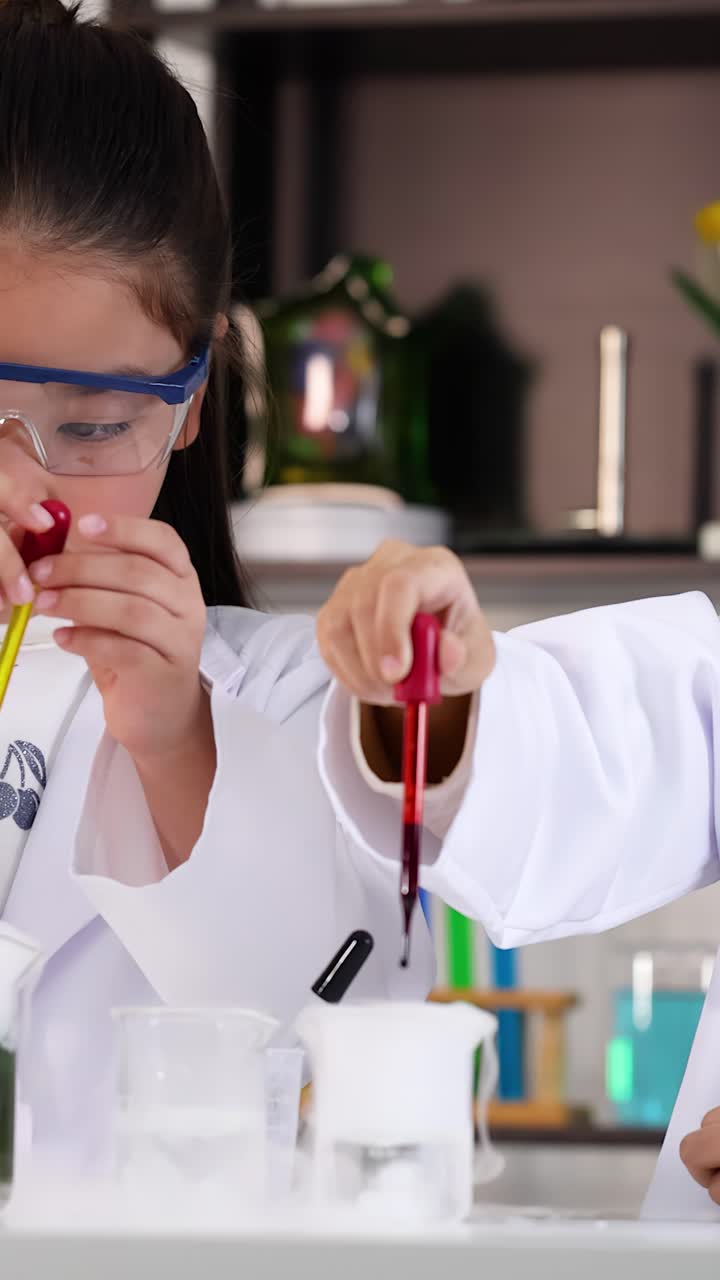 Dua gadis muda Asia berjas lab menggunakan pipet untuk eksperimen kimia yang penuh warna.