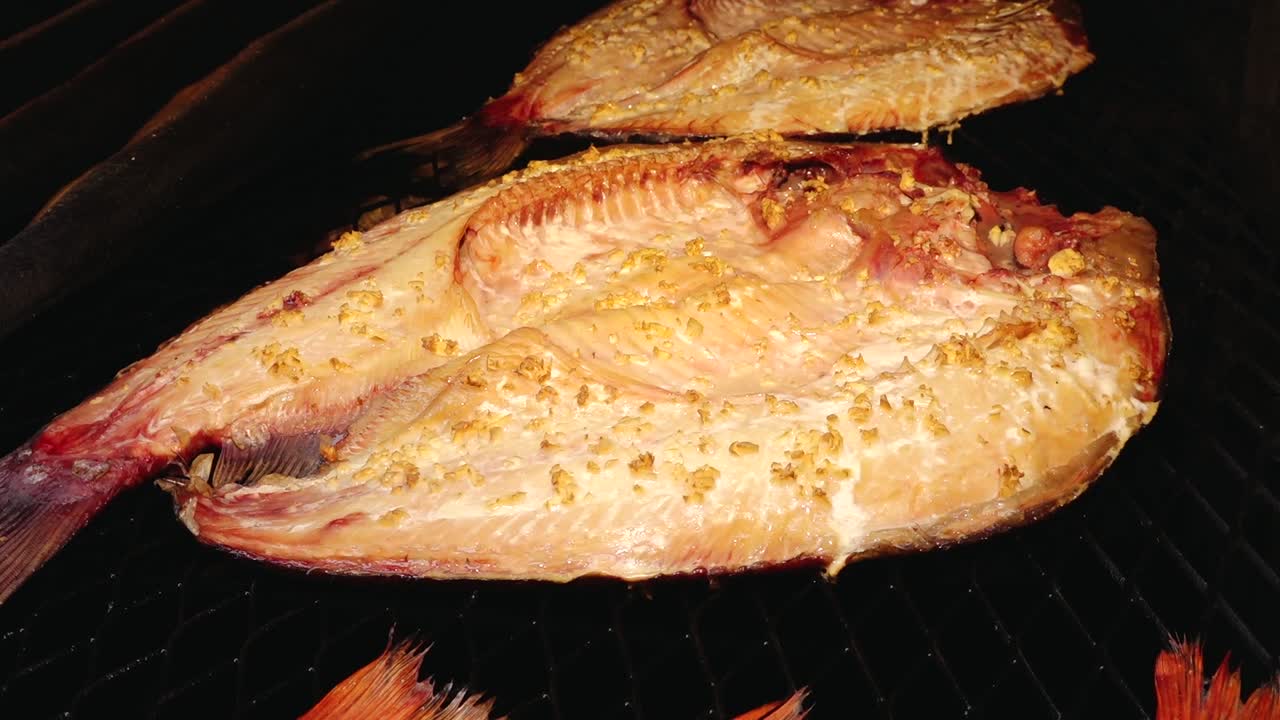 Various types of fish, including eel and redfish, are being smoked on a grill rack. The golden-brown color and rich texture highlight the traditional smoking process.