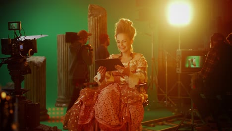 on period costume drama film set: beautiful smiling actress wearing renaissance dress, sitting on chair using digital tablet computer with green screen in background. high budget period drama. zoom in