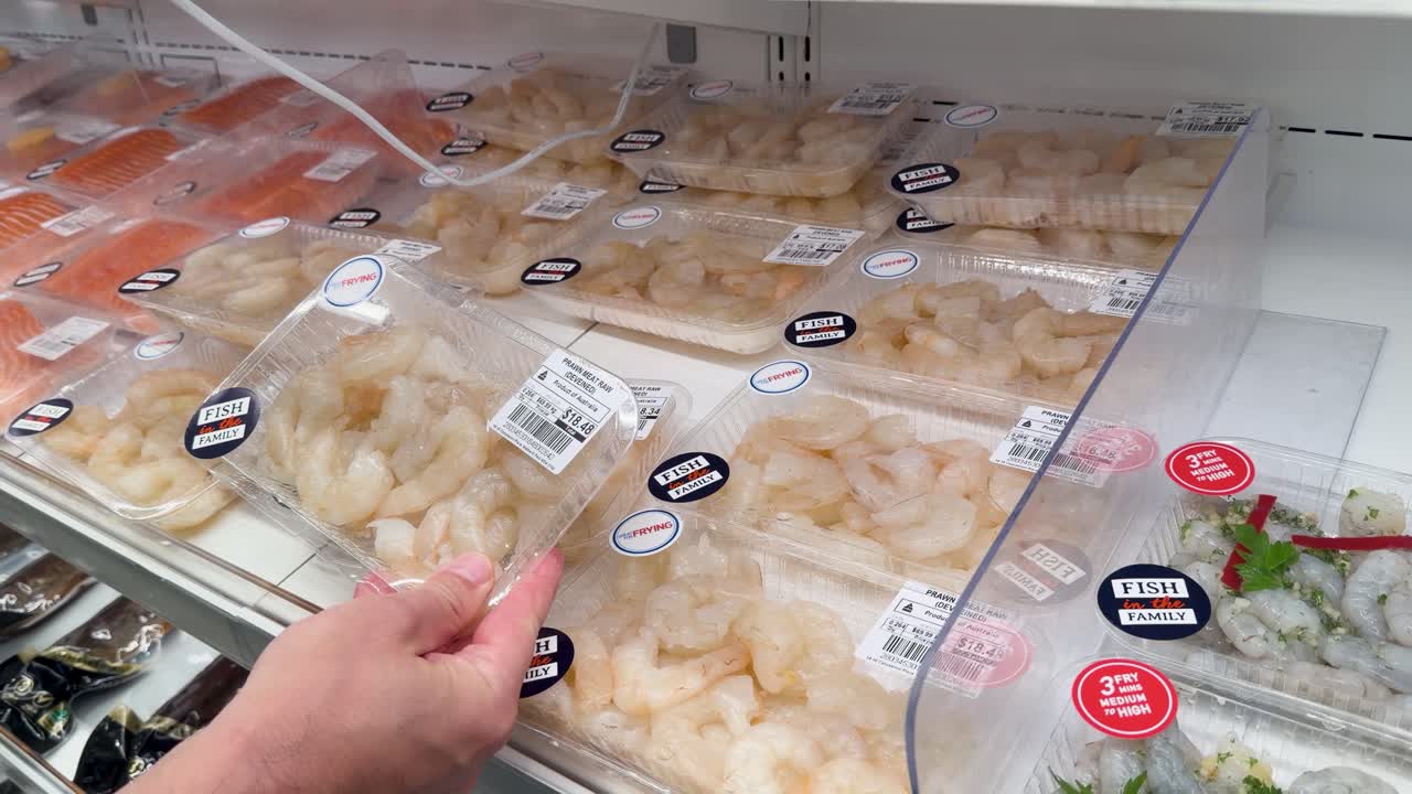 A hand reaches into a grocery store refrigerator to select a tray of peeled shrimp
