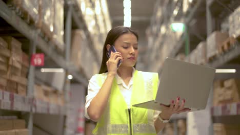 asian woman in uniform talks on mobile phone discuss amount of stock product inventory on shelf at distribution warehouse factory while work on laptop, logistic business shipping and delivery service