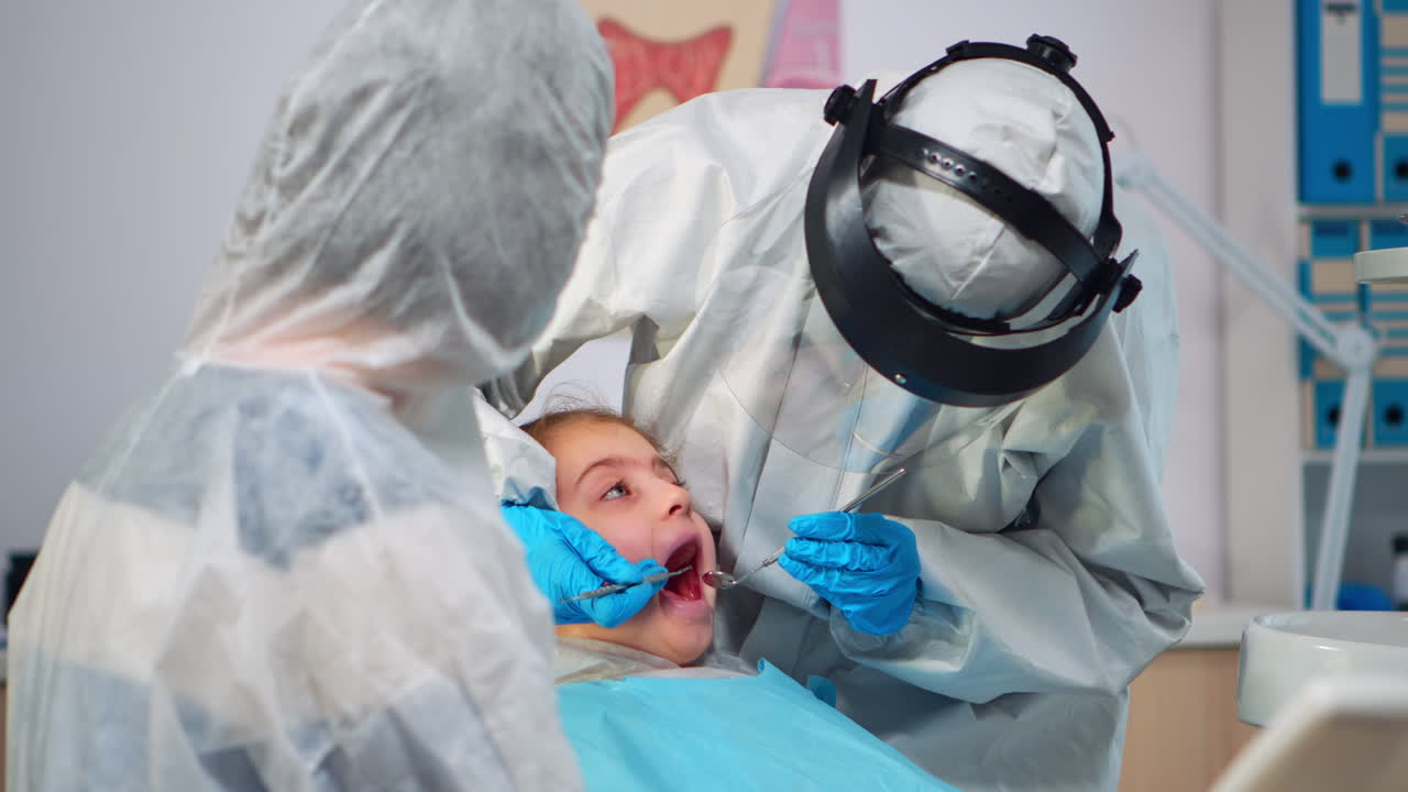 Primo piano di un dentista in tuta protettiva che esamina una paziente bambina in uno studio dentistico nella nuova normalità