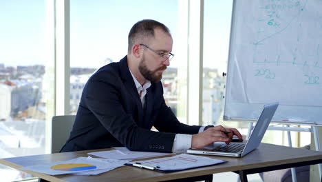 serious male worker using laptop computer. focused businessman working on laptop in office with large windows. businessman in glasses typing on a computer.