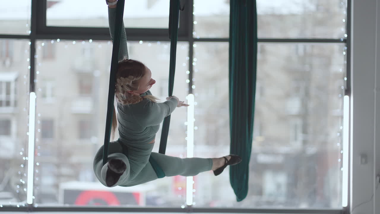 Una giovane donna sorridente si allena con un'amaca da aero stretching. Esegue esercizi di yoga aereo in sospensione in un'amaca verde in una palestra.