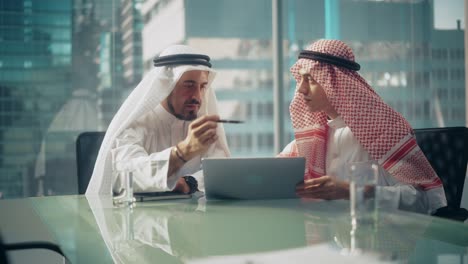 dos empresarios emiratíes exitosos con trajes tradicionales blancos sentados en una reunión de oficina, negociando y hablando sobre oportunidades de inversión, usando una computadora portátil. concepto de empresario saudí, emiratí, árabe.