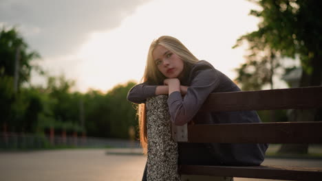 lady seated on bench at the edge, resting her chin on her arms, with sunlight creating reflective light around her and a backdrop of greenery, she appears calm and thoughtful