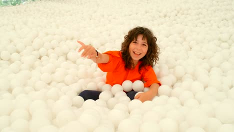 a cute 9-10 years old girl having fun among lots of white ball in a ball pit. 4k slow motion