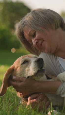 primer plano de una mujer acostada en un campo de hierba sosteniendo a su perro con afecto mientras acaricia su cara, bajo la cálida luz del sol, con un fondo borroso de árboles y vegetación