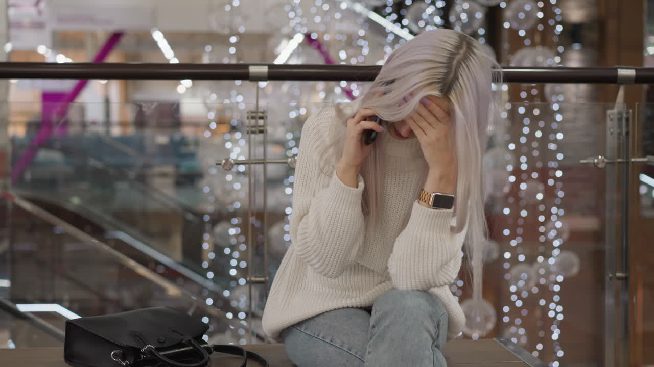Soft spoken lady sitting on bench in mall walkway, supporting chin with hand while talking on phone against backdrop of decorative lights and glass railing, casual outfit and purse beside her