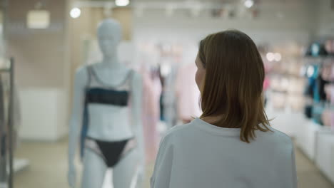 woman standing in front of retail store observing mannequin dressed in lingerie, contemplating while surrounded by blurred background with racks of clothing in well-lit mall environment