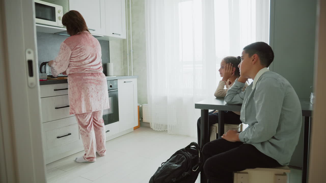 mother in pink velour pajamas preparing breakfast at kitchen countertop while children in school uniforms sit at table waiting with backpacks near window bright morning home routine scene