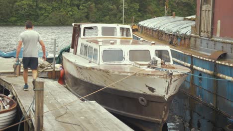 un joven carpintero camina por el embarcadero hasta un barco de madera con una lijadora de banda.