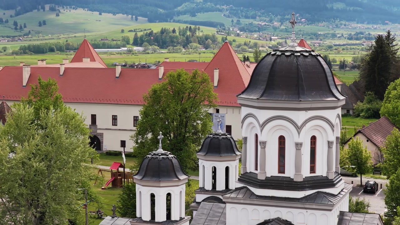 Vista aérea con dron de una iglesia ortodoxa con cúpulas negras junto a una fortaleza y colinas verdes, Harghita, Rumanía