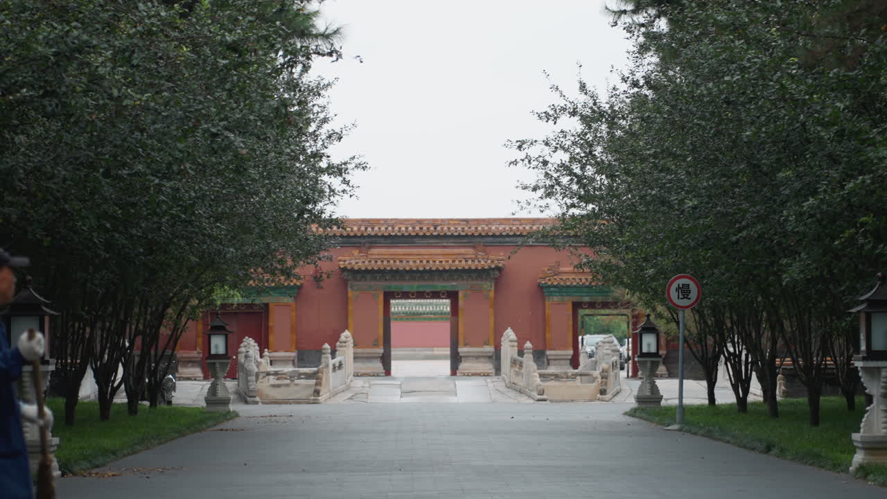 Cancello rosso del tempio incorniciato dagli alberi, un singolo visitatore in veste blu scuro cammina lungo un sentiero lastricato verso l'ingresso di pietra, cielo coperto, lanterne e siepi curate, l'architettura tradizionale evoca un pellegrinaggio sereno