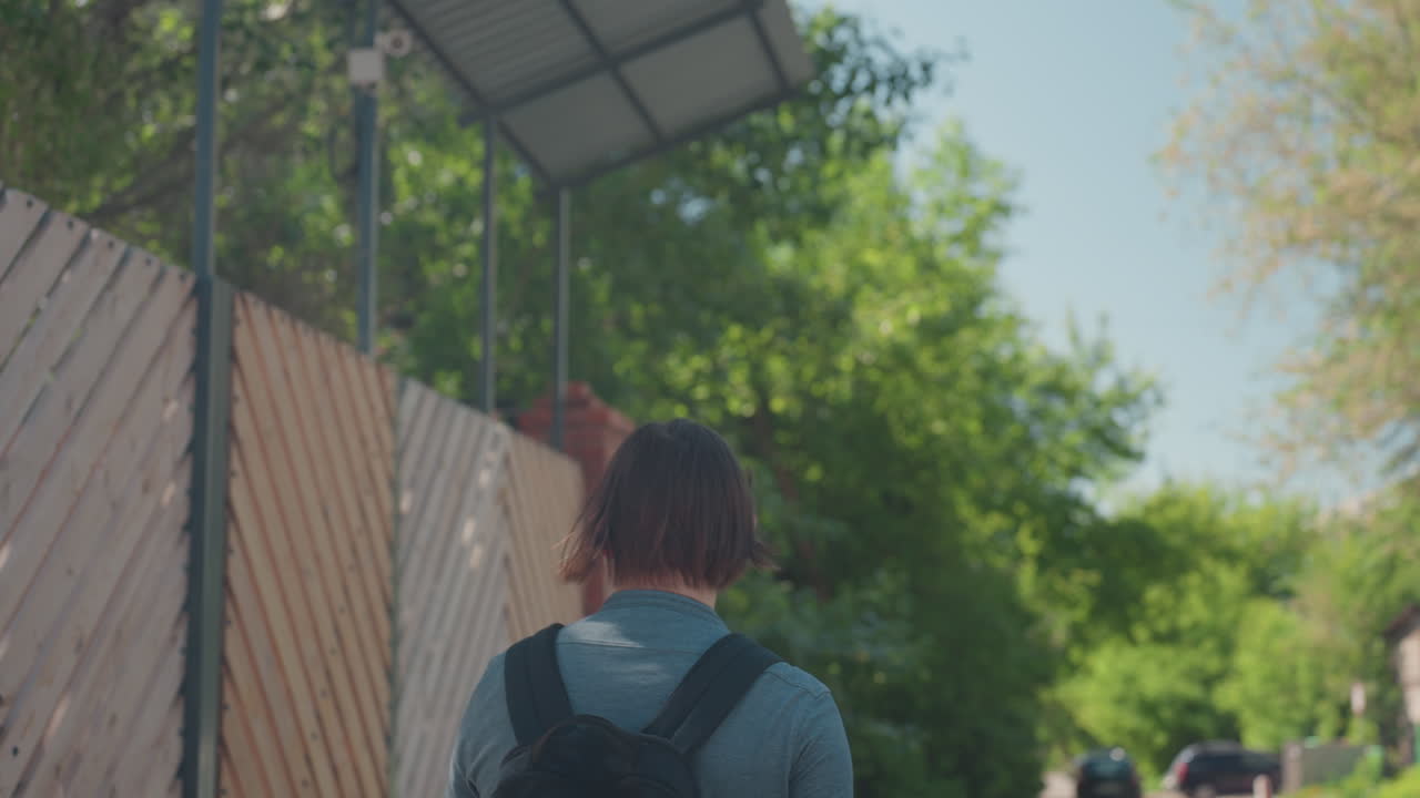 흰색 티셔츠를 입은 십 대 학생이 나무 울타리가 있는 교외 길을 백팩을 메고 걷고 있어요. 잎이 무성한 나무들이 인도를 따라 늘어서 있고, 따뜻한 오후 햇살이 내리쬐는 조용한 주택가에서 집으로 천천히 발걸음을 옮기는 모습이에요.