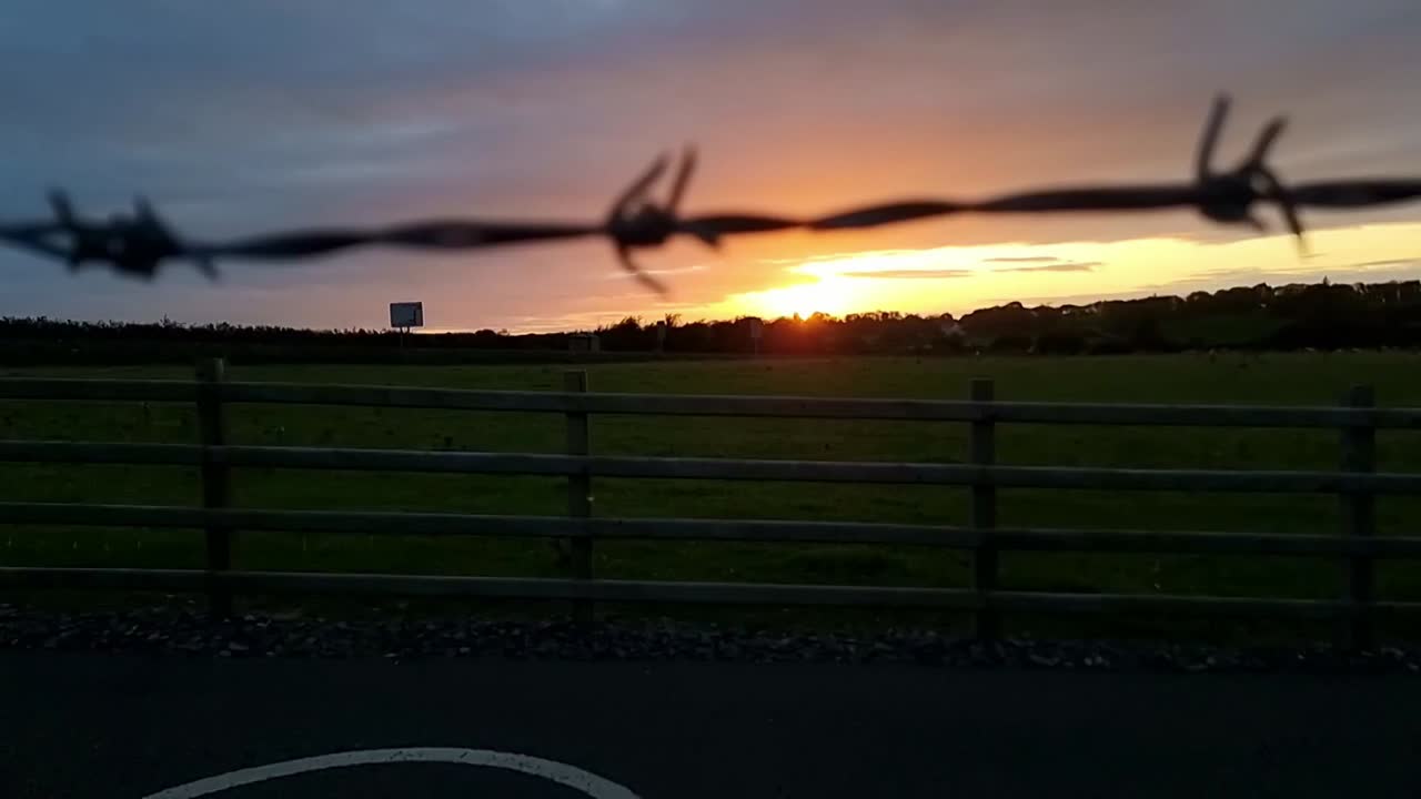 Looking through barb wire fence to sunset orange skyline over agricultural farmland field