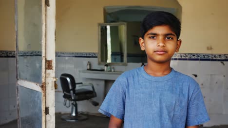 portrait of a young boy standing in his old abandoned barbershop waiting for customers, wearing a blue t shirt, with a serious expression on his face