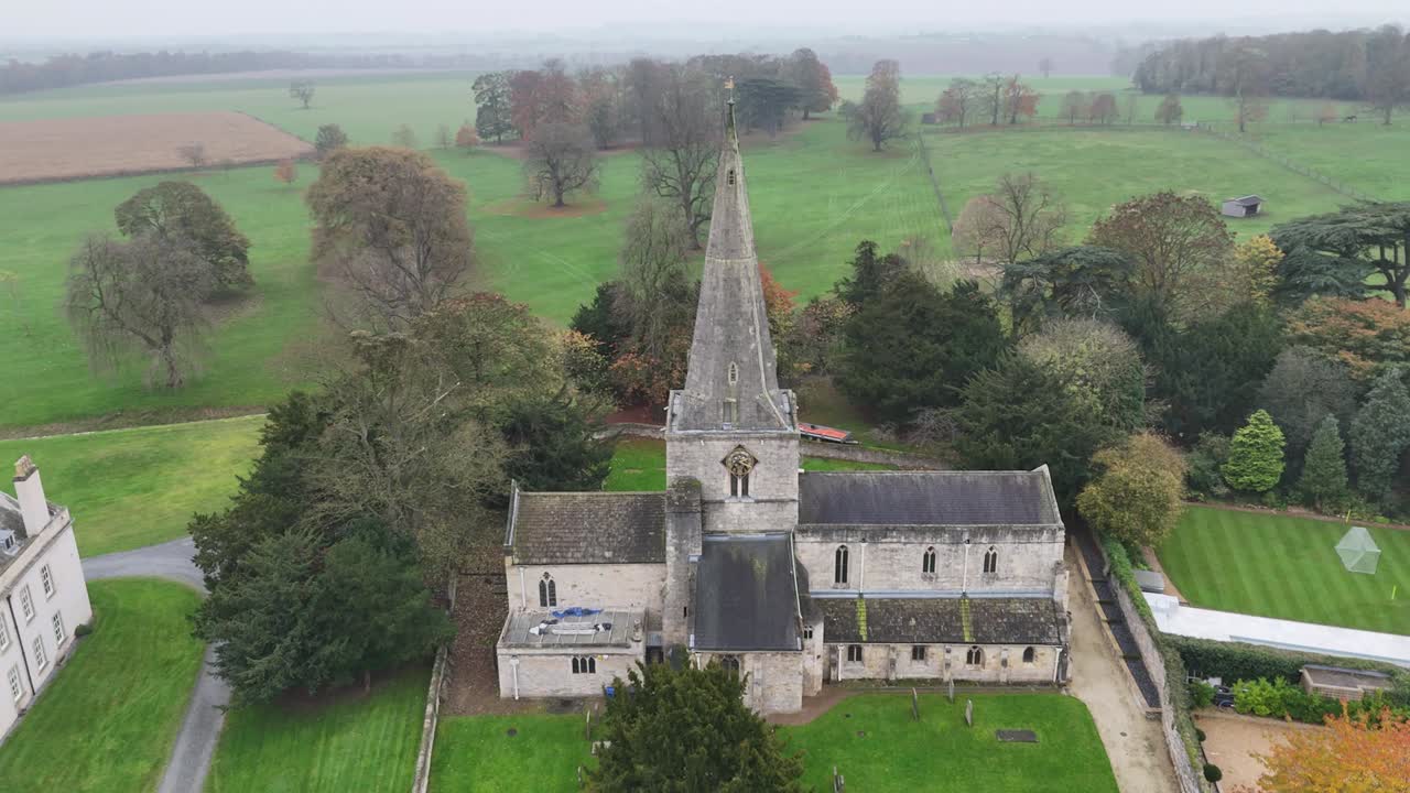 An aerial shot reveals St Peter's Church in Cassington, Oxfordshire, a historic English church with a prominent spire, nestled amidst green fields and trees.