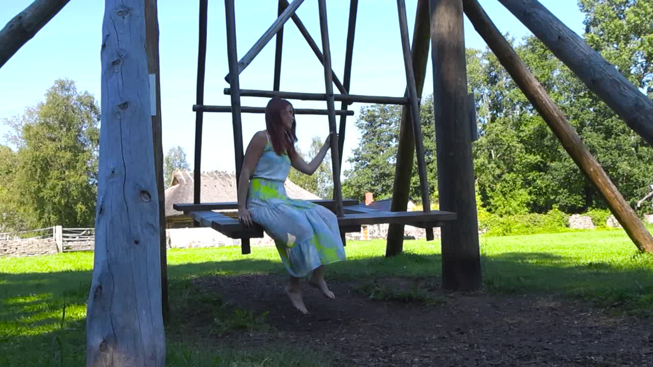Gorgeous woman with red hair and long white skirt or dress swinging on a traditional wooden swing in an open air museum with farm houses and grassy green garden around her during summer sunny day